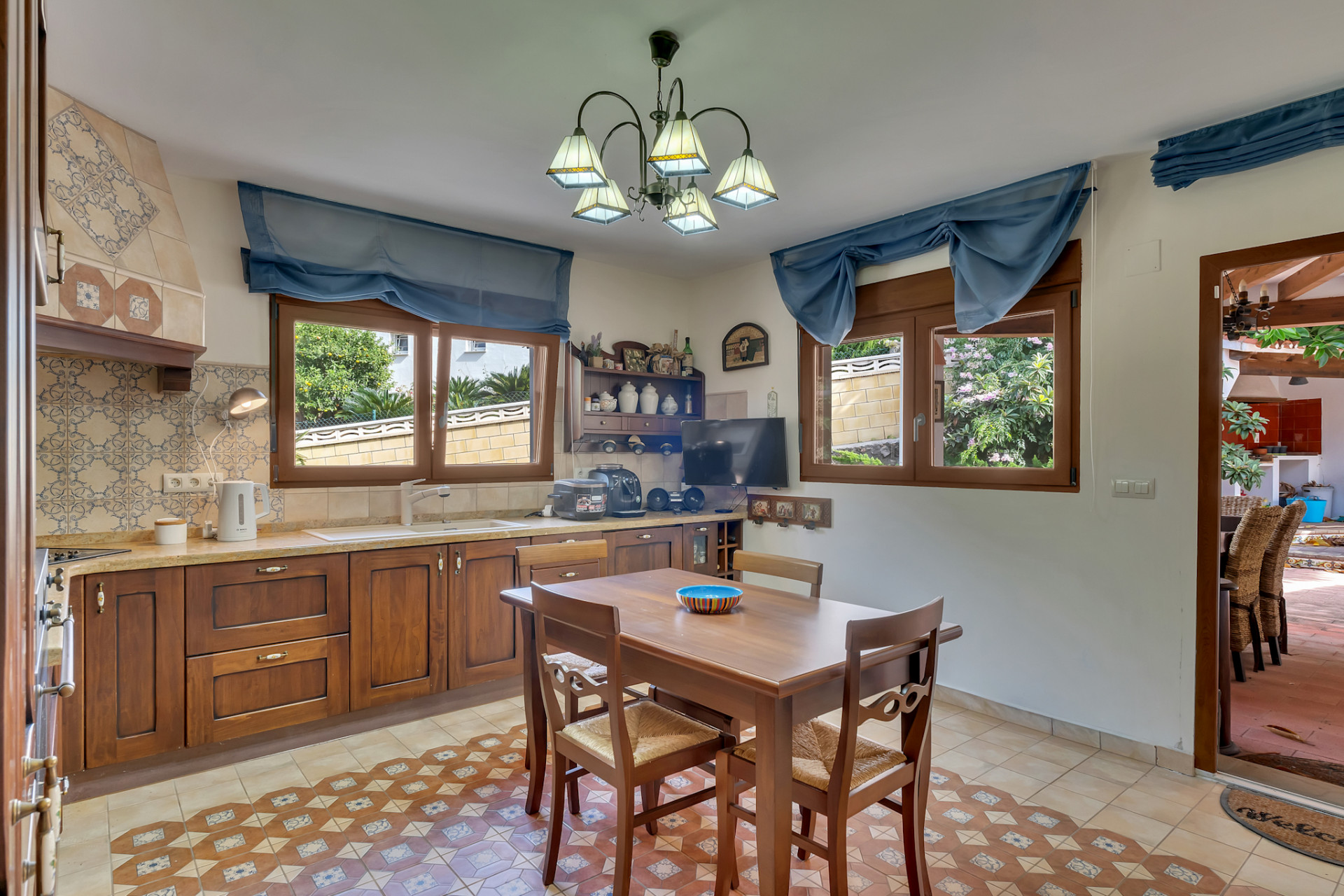 Functional kitchen featuring dark wood cabinetry, decorative tiled backsplash, and patterned floor tiles. Two windows offer garden views, complemented by a wooden dining table.
