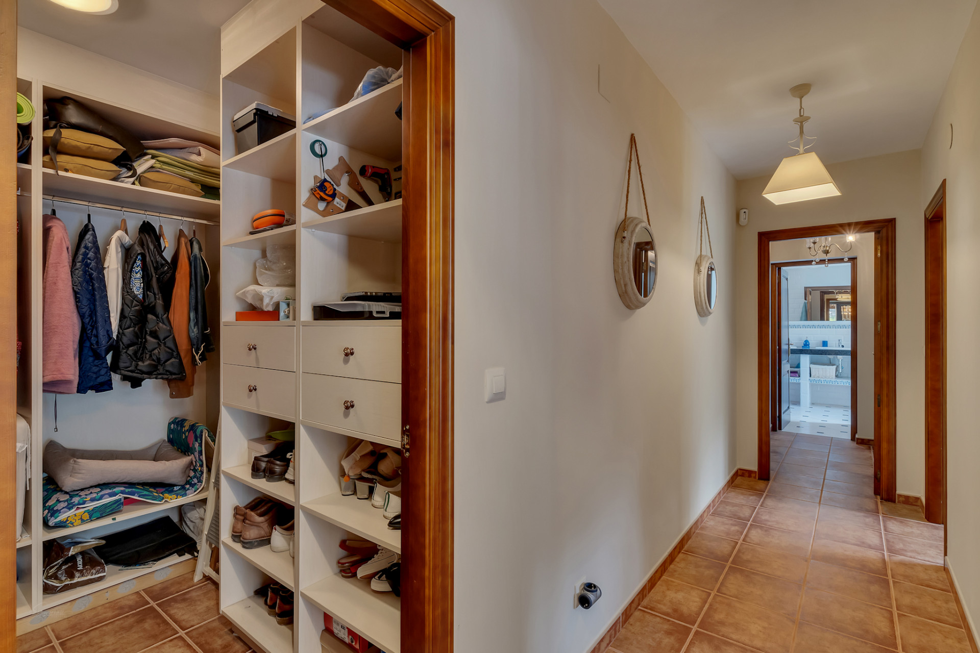 Hallway with an integrated open wardrobe featuring white shelving and drawers, terracotta floor tiles, and wooden door frames.