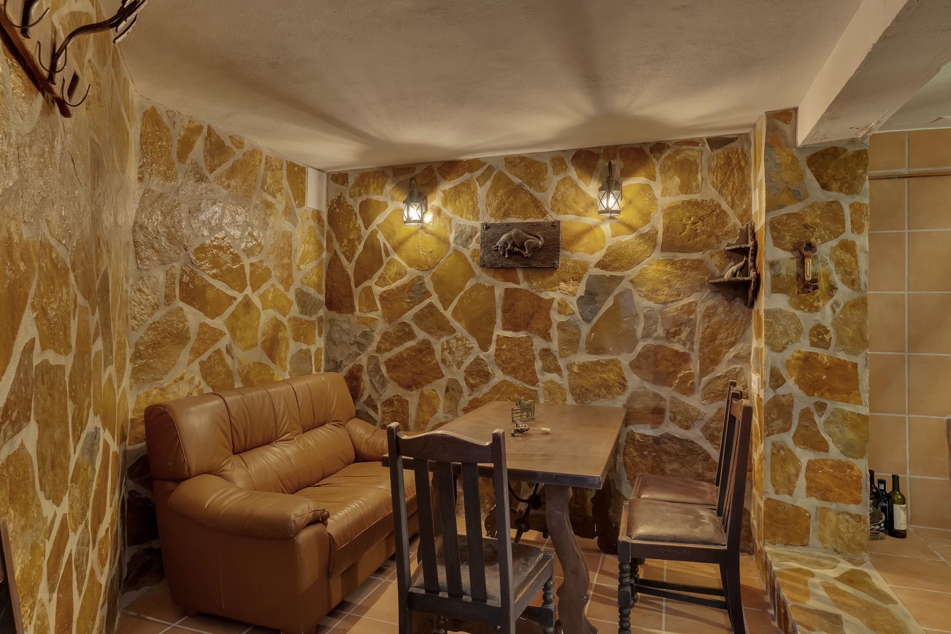 Interior space featuring irregular stone-clad walls and tiled flooring. Furnished with a leather sofa, rustic wooden table, and warm lantern wall lights.