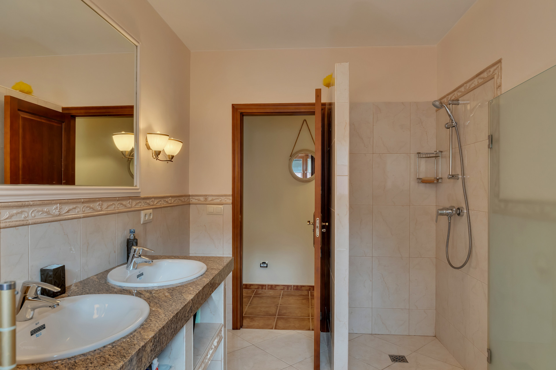 Main bathroom featuring a double vanity with granite countertop, a large framed mirror, and a walk-in shower with glass partition, complemented by warm-toned tiling.