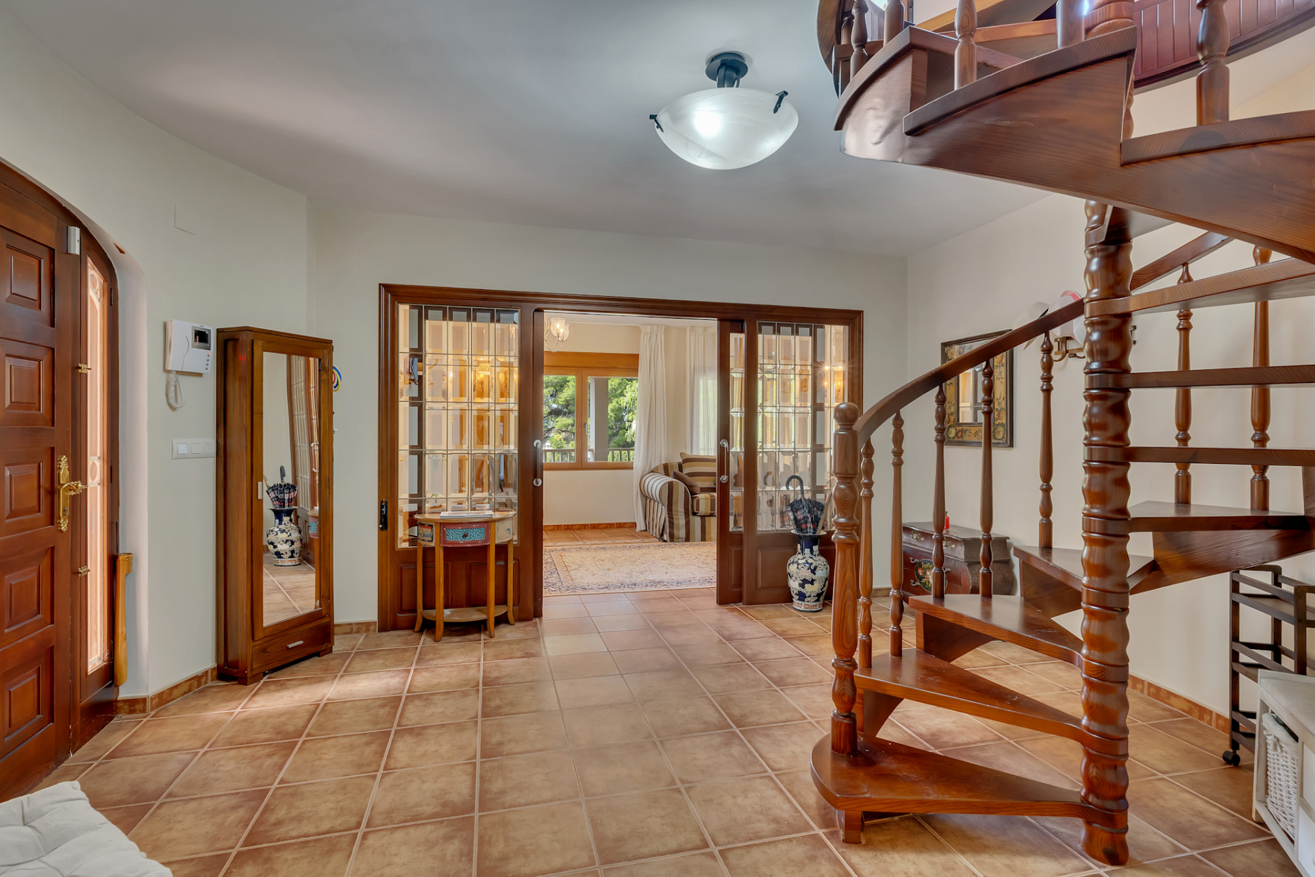 Main entrance featuring a dark wooden spiral staircase and terracotta tiled flooring. Wooden and glass sliding doors open to a bright adjoining room.
