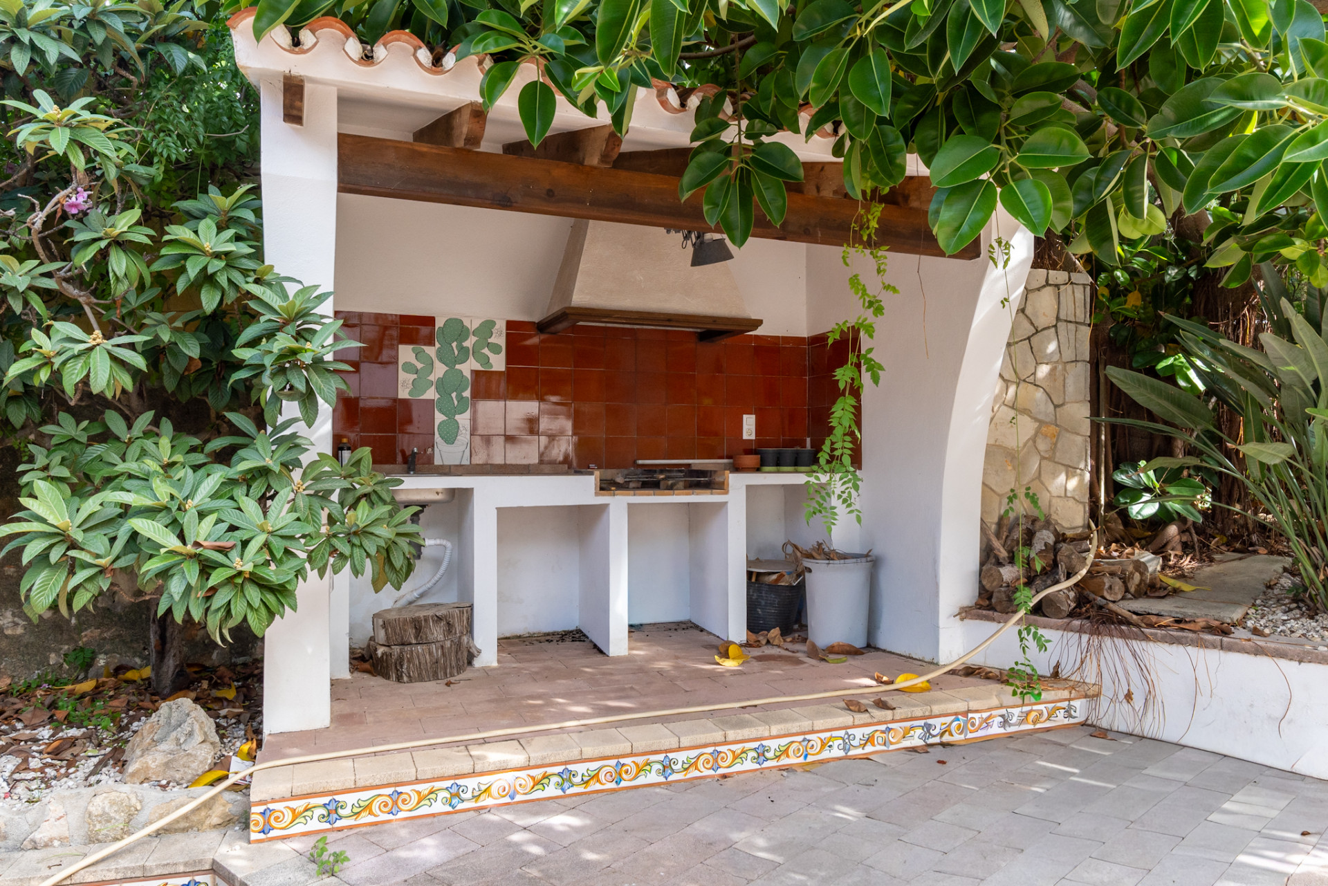 Outdoor kitchen with a built-in BBQ grill, terracotta tiled backsplash with a cactus motif, and a rustic wooden beam ceiling. The area features a tiled floor and is framed by lush greenery.