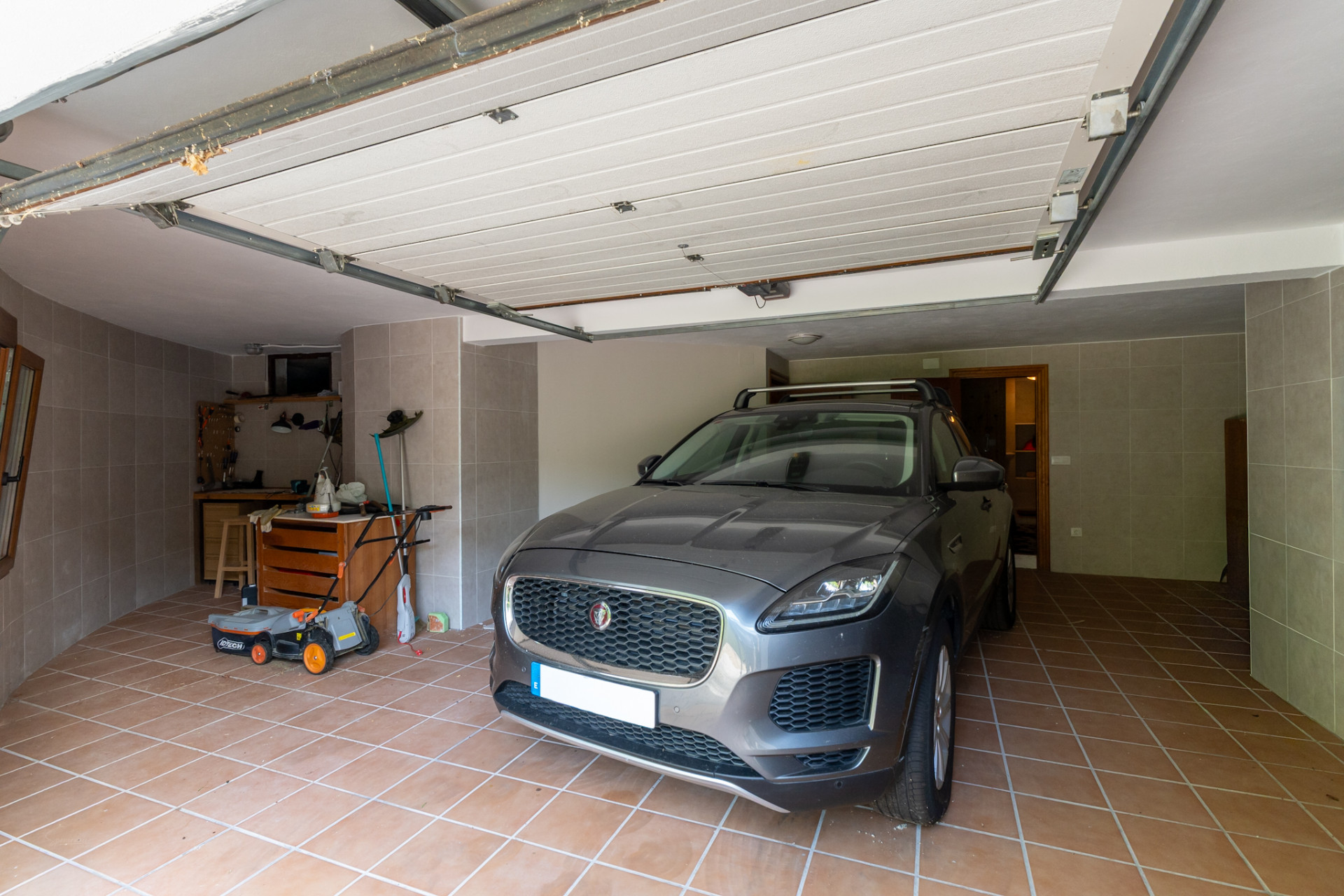 Private garage featuring terracotta tiled flooring and grey tiled walls. It includes a dedicated workbench area with tools, a lawnmower, and an open sectional door.