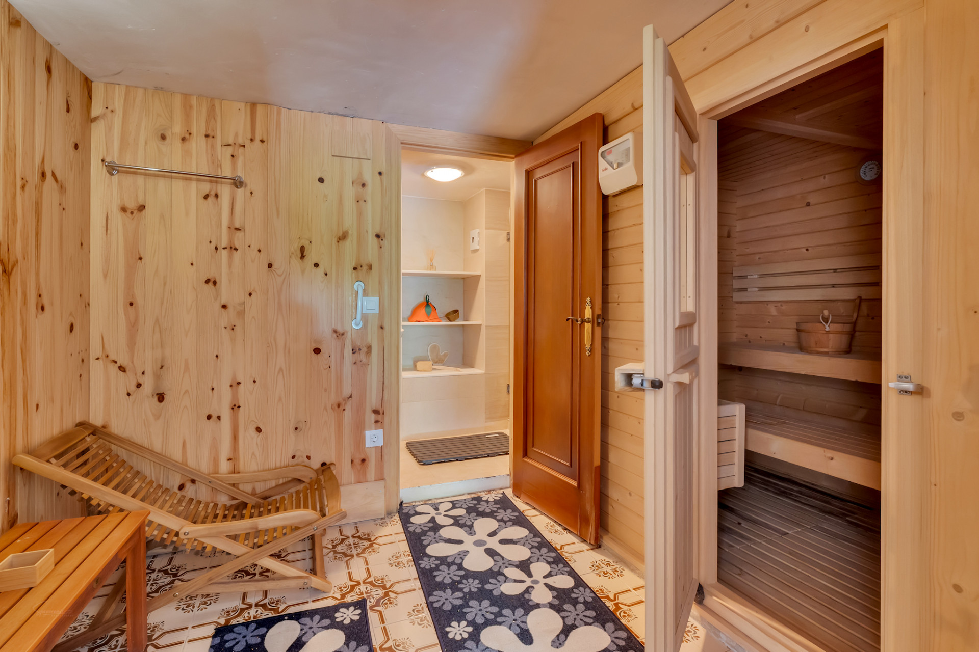 Relaxation area featuring light wood-paneled walls, tiled flooring, and direct access to a pine wood sauna. Includes a wooden lounge chair and an adjacent utility space with shelving.