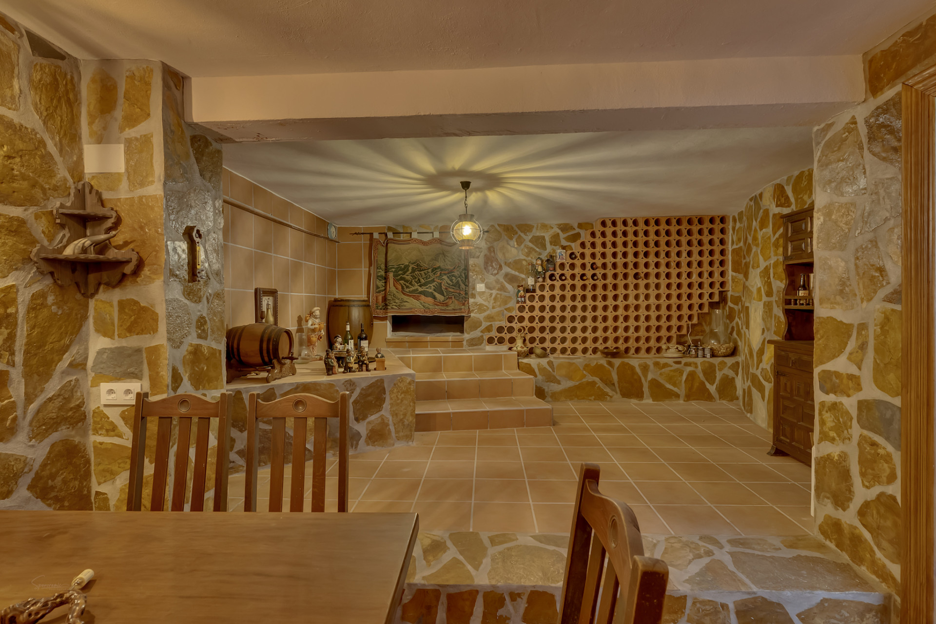 Rustic cellar featuring natural stone walls, terracotta floor tiles, and a prominent built-in wine rack. A central light fixture creates a unique ceiling pattern, complementing the traditional decor and wooden elements.