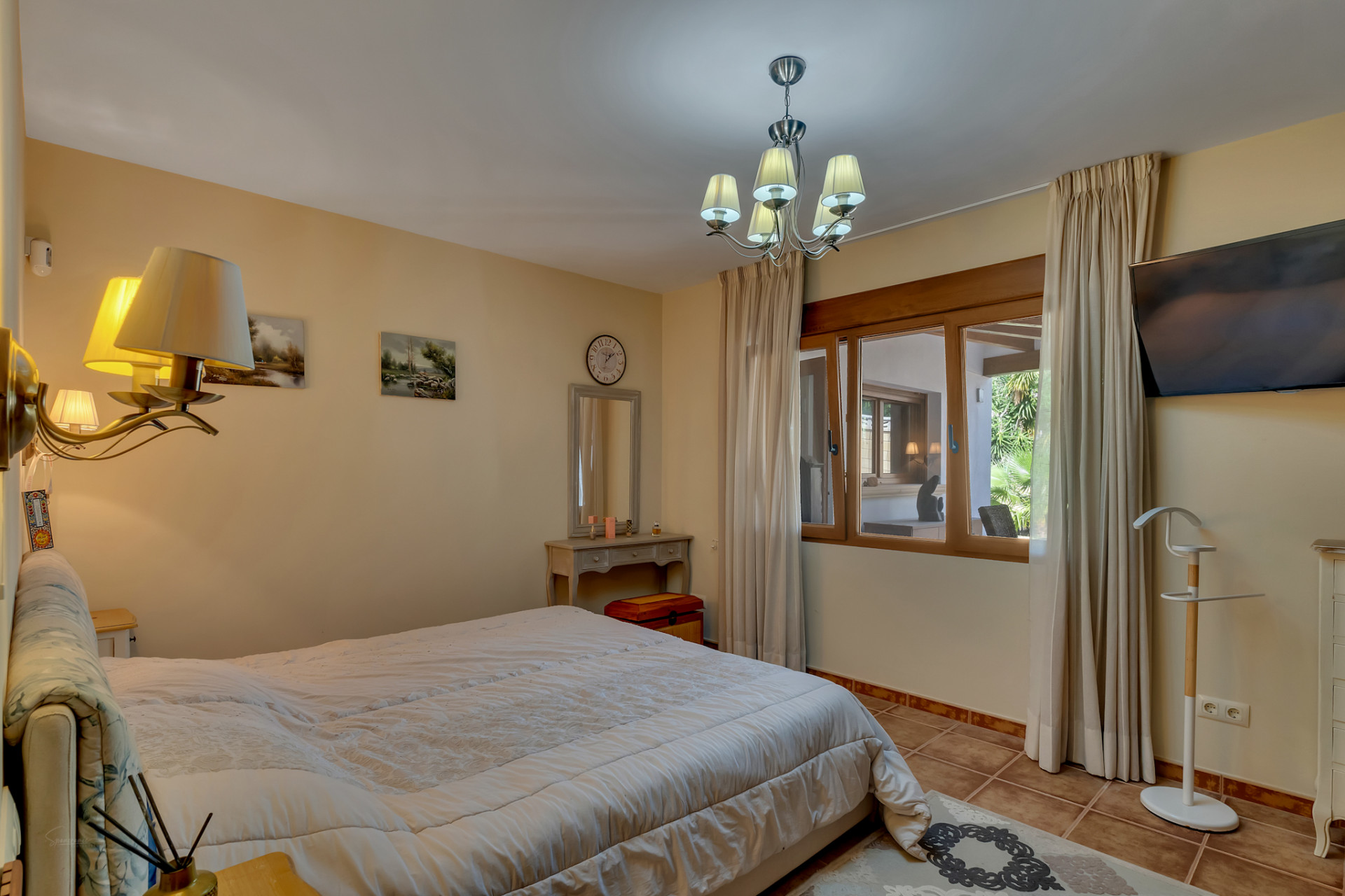 Spacious bedroom featuring a large bed, tiled floor, and a wooden-framed window offering views of an adjacent covered area and garden. Includes a dressing table and TV.