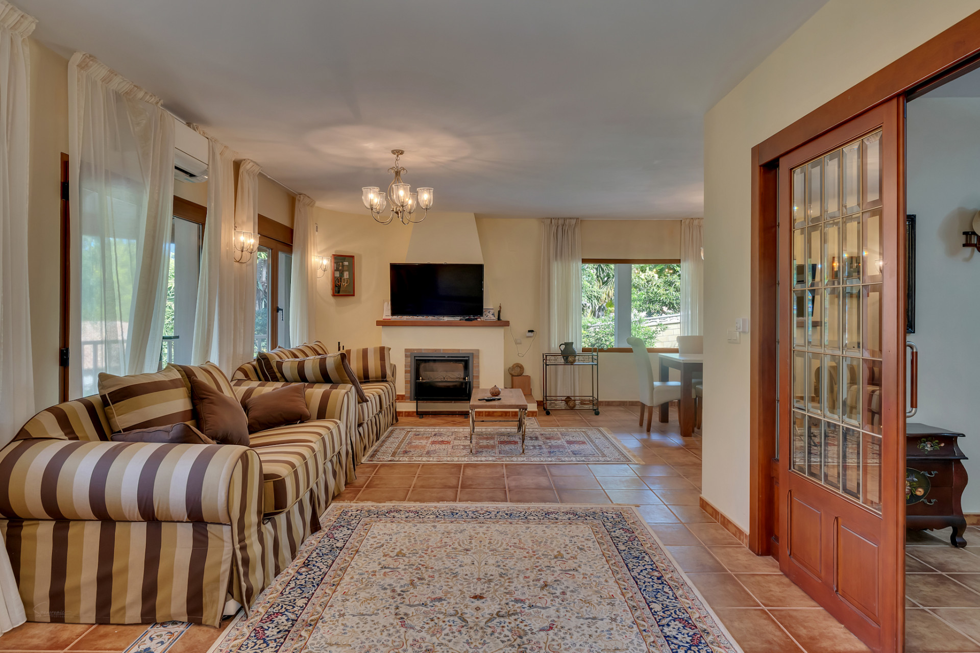 Spacious living room featuring a striped sofa, fireplace, and television. Terracotta tiled floor, large windows with sheer curtains, and a wooden sliding glass door.