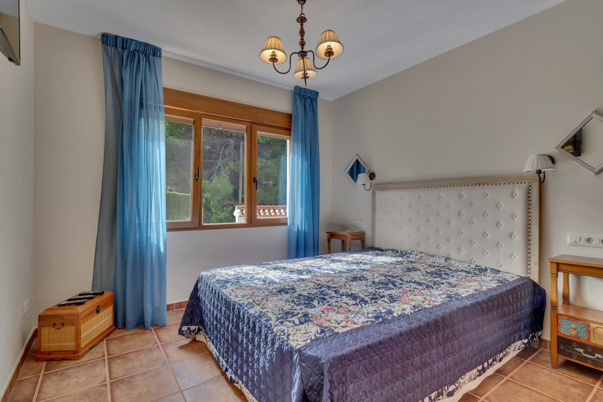This bedroom features a large bed with a white tufted headboard, a blue patterned bedspread, and terracotta-tiled floors. A wooden-framed window with sheer blue curtains provides natural light and views of lush greenery.