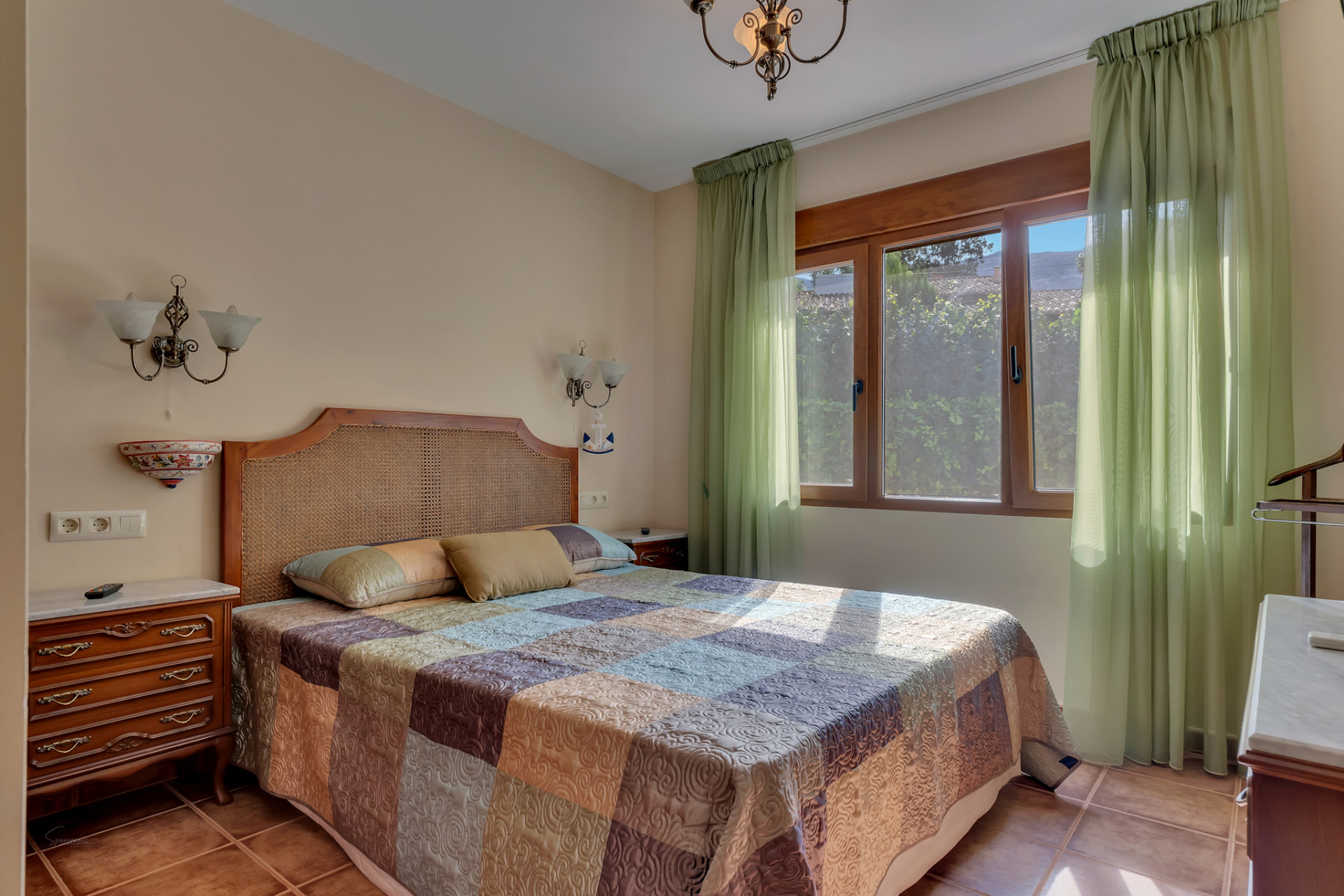 This bright bedroom features a rattan headboard, a patterned quilt, and a large wooden-framed window with sheer green curtains. Terracotta floor tiles and classic wooden bedside tables complete the space.