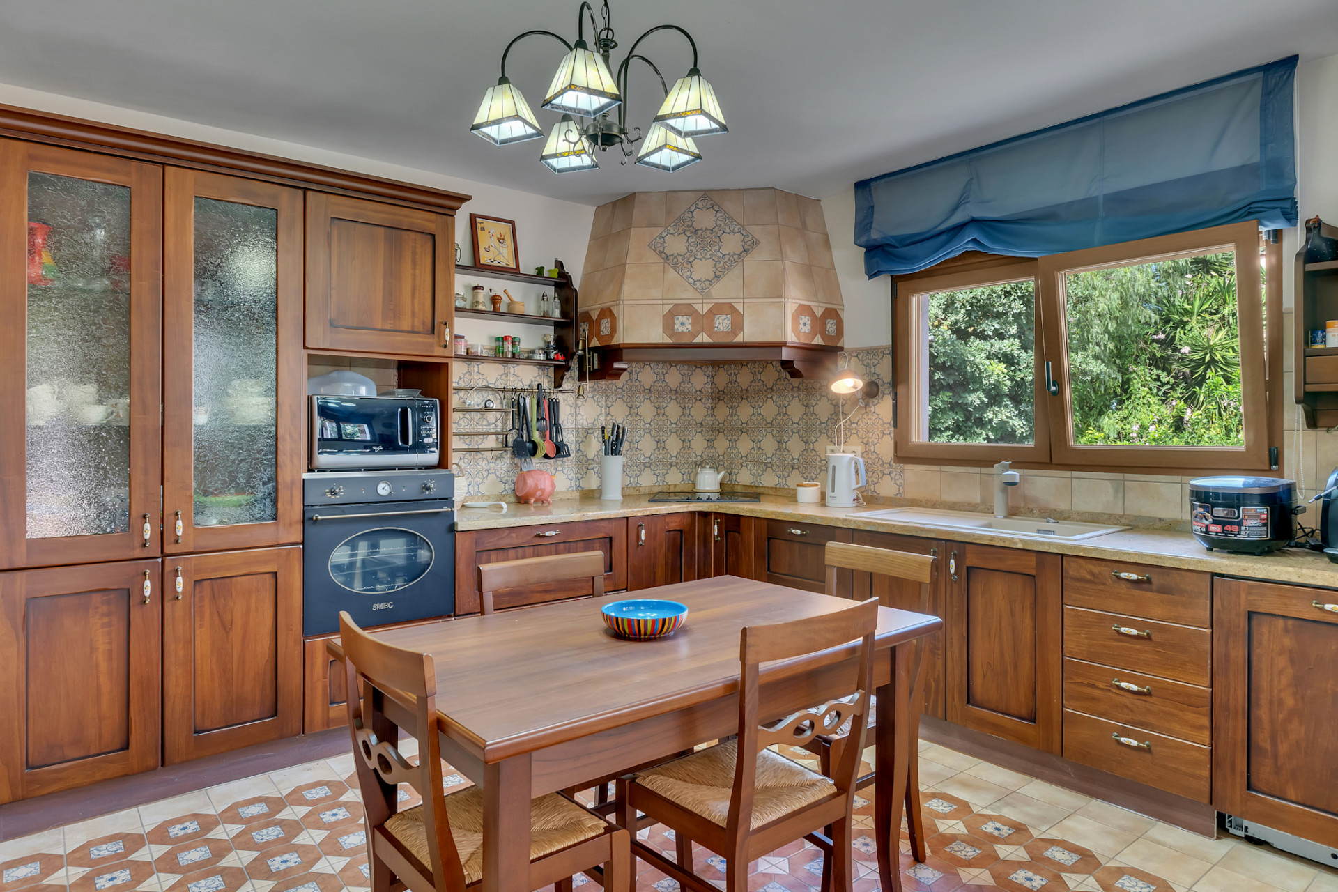 Traditional kitchen featuring dark wood cabinetry, a tiled backsplash with intricate patterns, and a central dining table. Natural light streams from the window overlooking lush greenery.