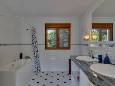 Bathroom featuring a double vanity with dark countertop, two sinks, and a large mirror. White subway tiles with a blue decorative border complement the patterned floor and wooden window frame overlooking greenery.