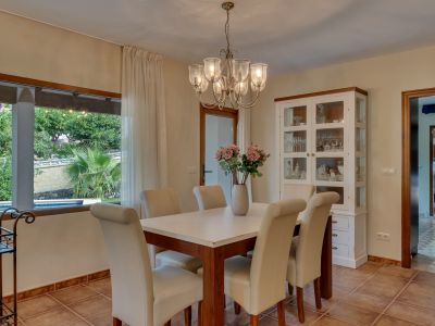 Bright dining room with a wooden table, cream upholstered chairs, and a chandelier. A large window offers views of the lush garden and private pool.