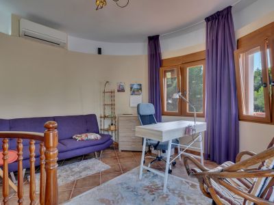 Bright office space featuring a purple sofa, light wood desk with an ergonomic chair, and large wooden windows offering views of lush greenery. Includes an air conditioning unit and terracotta floor tiles.
