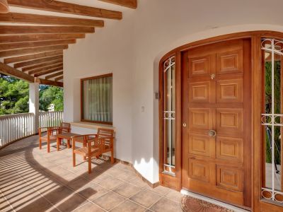 Covered entrance terrace with a paneled wooden door, tiled floor, and exposed wooden beams. Features a seating area and a curved railing overlooking lush greenery.