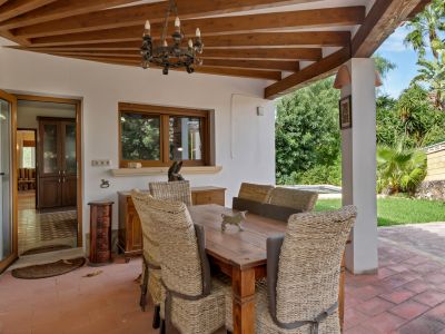 Covered outdoor dining area with a wooden beamed ceiling, terracotta floor, and a rustic chandelier. Wicker chairs surround a robust wooden table, opening to a lush garden and interior.