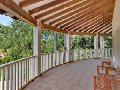 Curved covered terrace featuring a tiled floor and an exposed wooden beam ceiling. A white balustrade frames views of lush greenery, complemented by simple wooden seating.