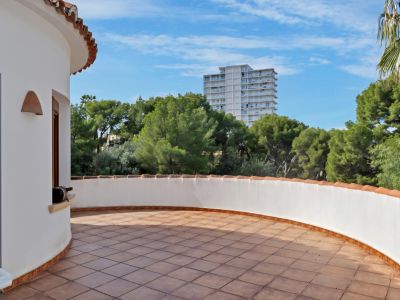 Curved outdoor terrace featuring terracotta-tiled flooring and a low white wall topped with traditional roof tiles. Surrounded by green trees with a distant building under a blue sky.