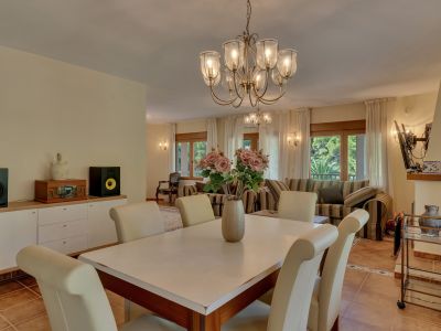 Elegant dining area featuring a classic chandelier and cream-colored chairs, seamlessly connecting to a bright living space with striped sofas and large windows overlooking lush greenery.