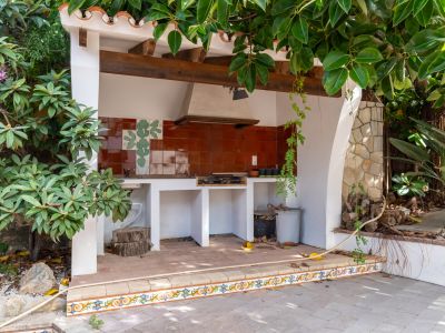 Outdoor kitchen with a built-in BBQ grill, terracotta tiled backsplash with a cactus motif, and a rustic wooden beam ceiling. The area features a tiled floor and is framed by lush greenery.