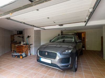 Private garage featuring terracotta tiled flooring and grey tiled walls. It includes a dedicated workbench area with tools, a lawnmower, and an open sectional door.