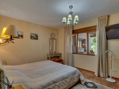 Spacious bedroom featuring a large bed, tiled floor, and a wooden-framed window offering views of an adjacent covered area and garden. Includes a dressing table and TV.