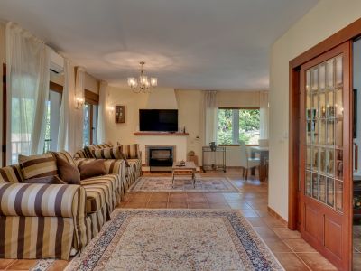 Spacious living room featuring a striped sofa, fireplace, and television. Terracotta tiled floor, large windows with sheer curtains, and a wooden sliding glass door.