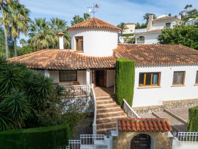 The villa's main entrance features a tiled staircase leading to a covered porch. White stucco walls, traditional terracotta roof tiles, and a distinctive circular tower characterize the exterior.