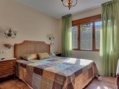 This bright bedroom features a rattan headboard, a patterned quilt, and a large wooden-framed window with sheer green curtains. Terracotta floor tiles and classic wooden bedside tables complete the space.