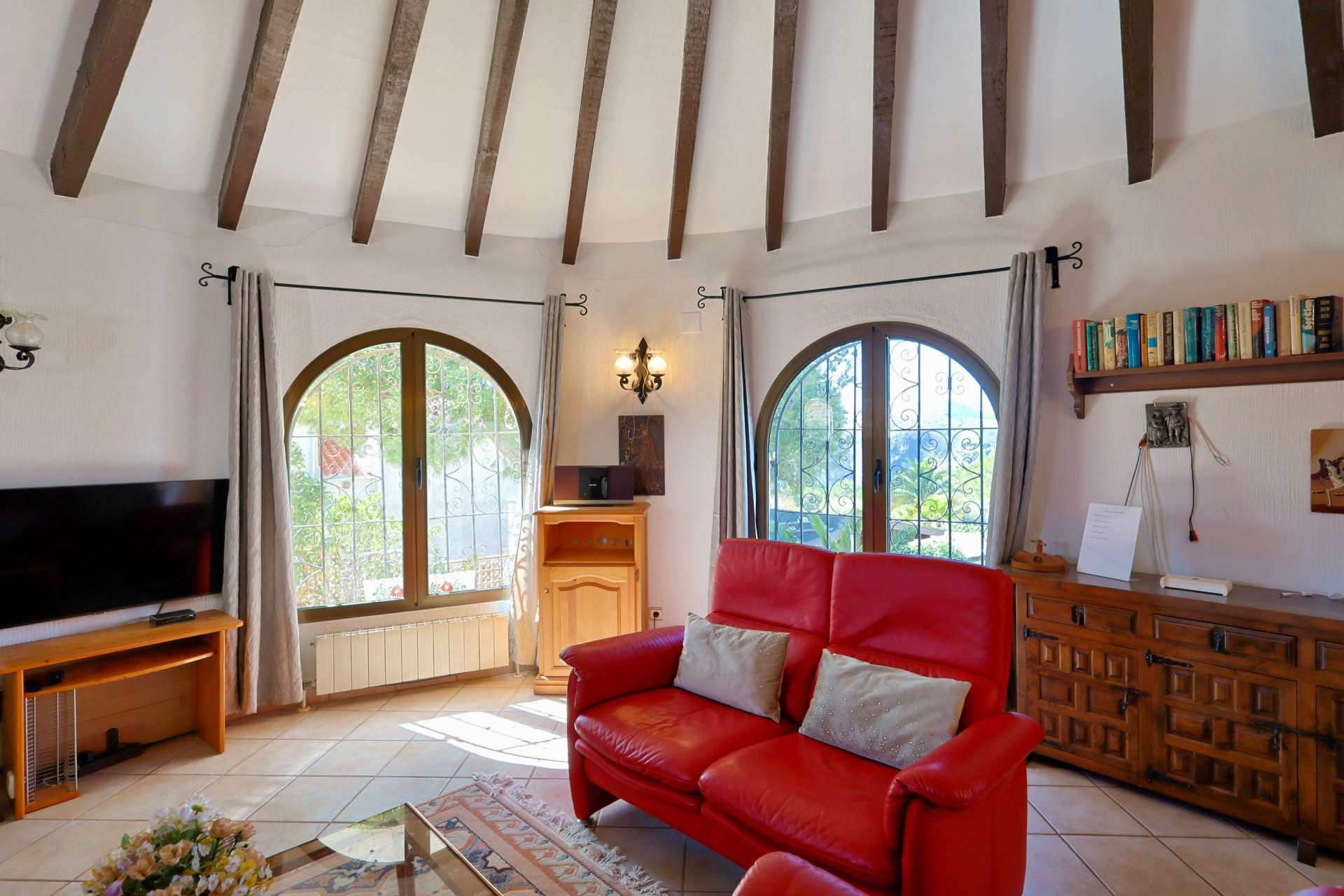 Bright living room featuring wood beams, a red leather sofa, and arched windows overlooking the garden.