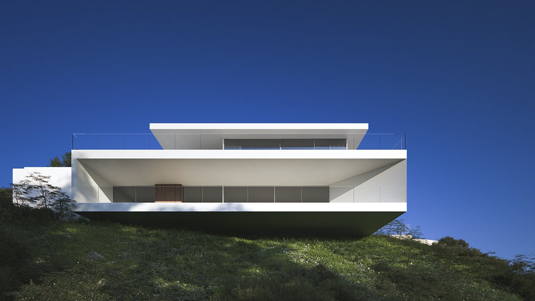 Exterior facade featuring minimalist white geometric cantilevers and glass railings, integrated into a natural hillside under a clear blue sky.