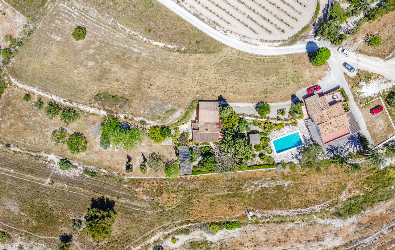 Aerial perspective of the estate showcasing the main villa, guest house, and central swimming pool area surrounded by lush gardens and agricultural plots.