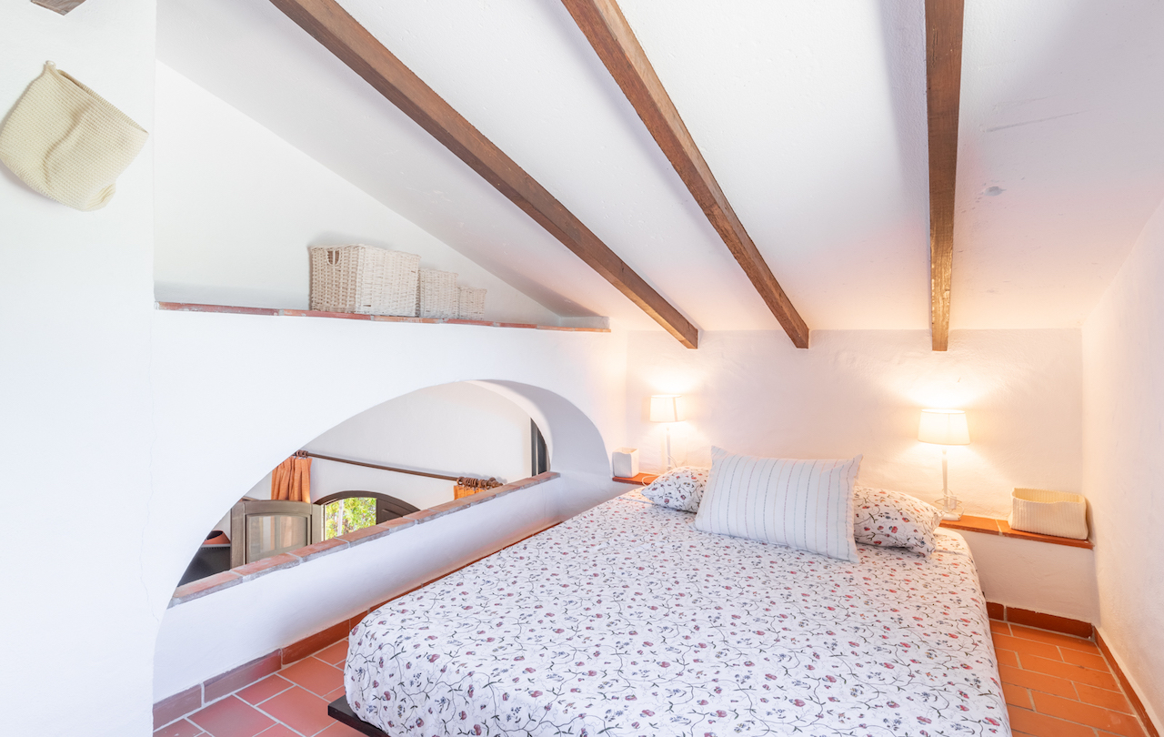 Attic bedroom featuring exposed wooden ceiling beams, terracotta tile flooring, and a unique arched opening overlooking the lower living area.
