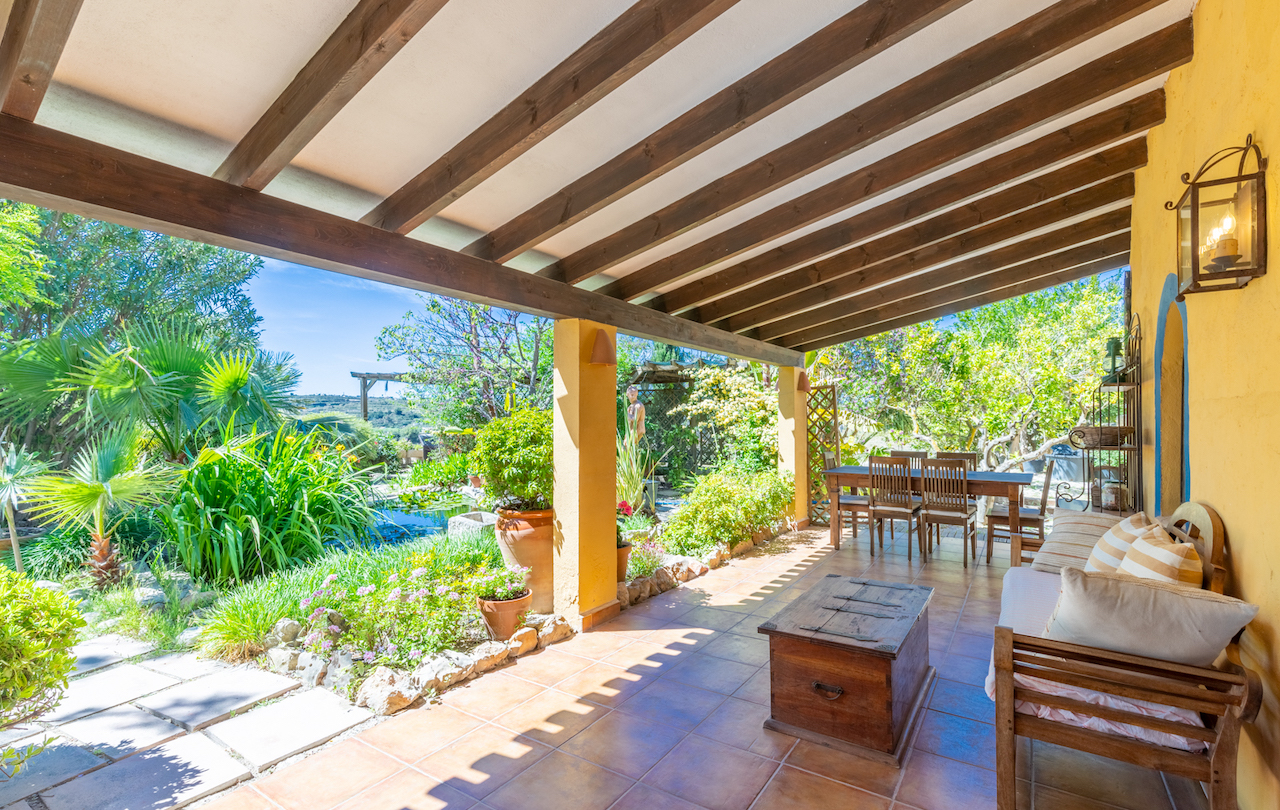 Covered terrace featuring exposed wooden beams, terracotta flooring, and outdoor living areas overlooking a lush Mediterranean garden with a pond.