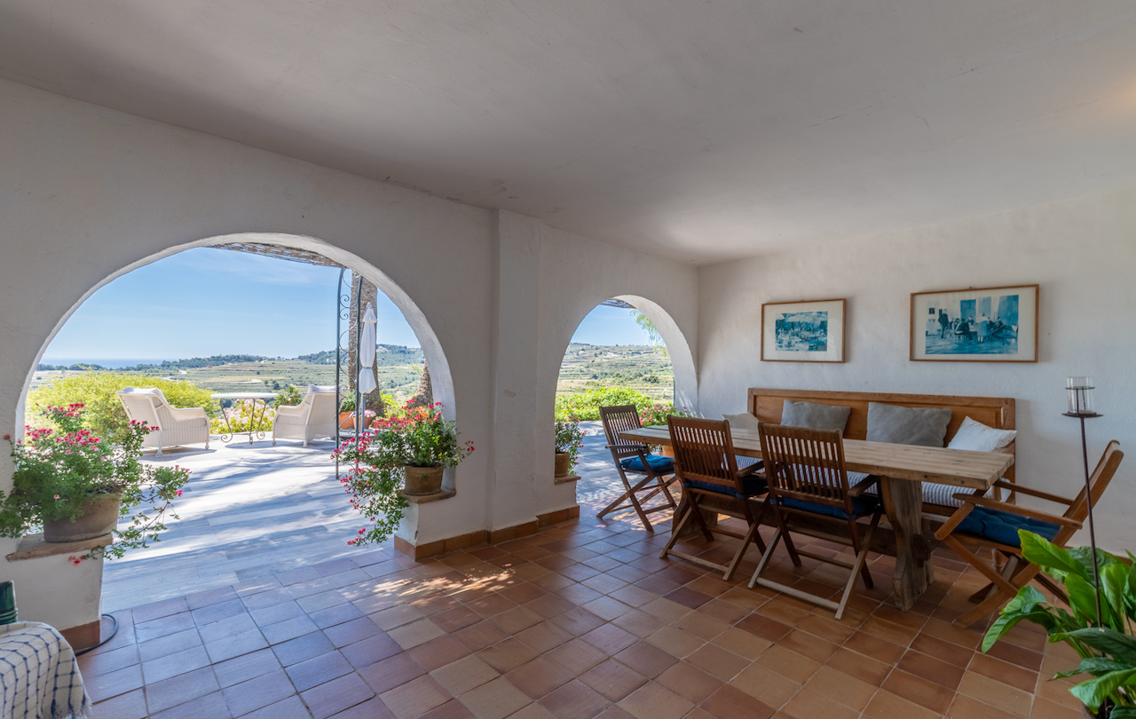 Covered terrace featuring traditional arches, terracotta flooring, and a rustic dining area. It overlooks the vineyards and the Mediterranean Sea from an elevated position.