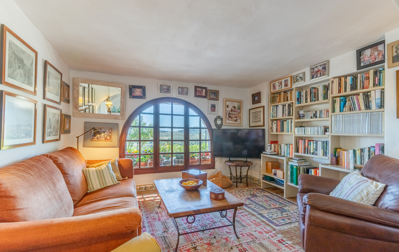 Living room library featuring a large arched wooden window, built-in shelving, terracotta flooring with rugs, and panoramic views of the surrounding landscape.