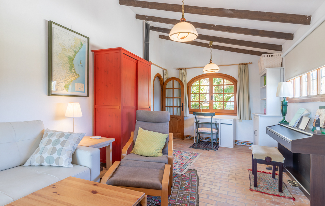 Living space featuring exposed wooden beams, terracotta floors, and a red wardrobe. Includes a workspace by an arched window and a digital piano.