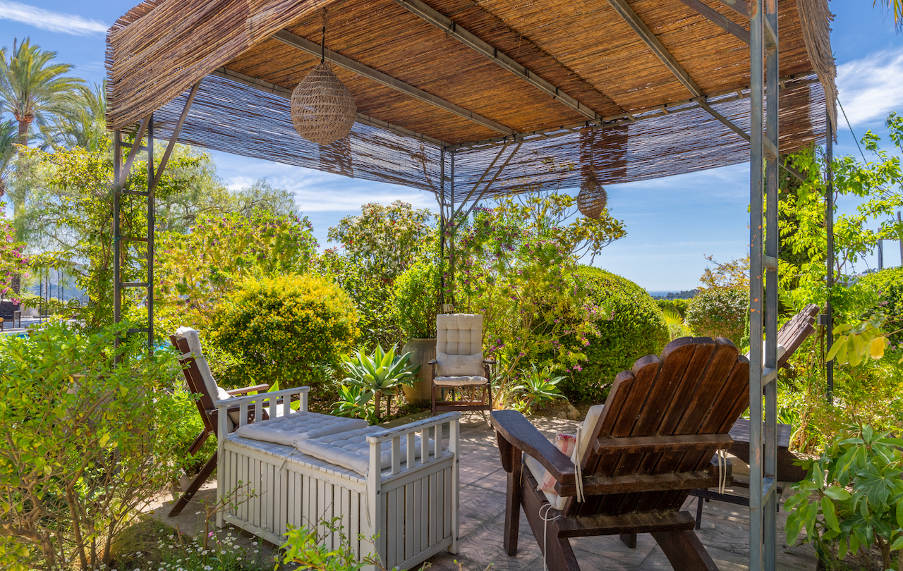 Metal-framed pergola with a reed roof featuring a lounge area, surrounded by lush Mediterranean gardens and distant sea views.