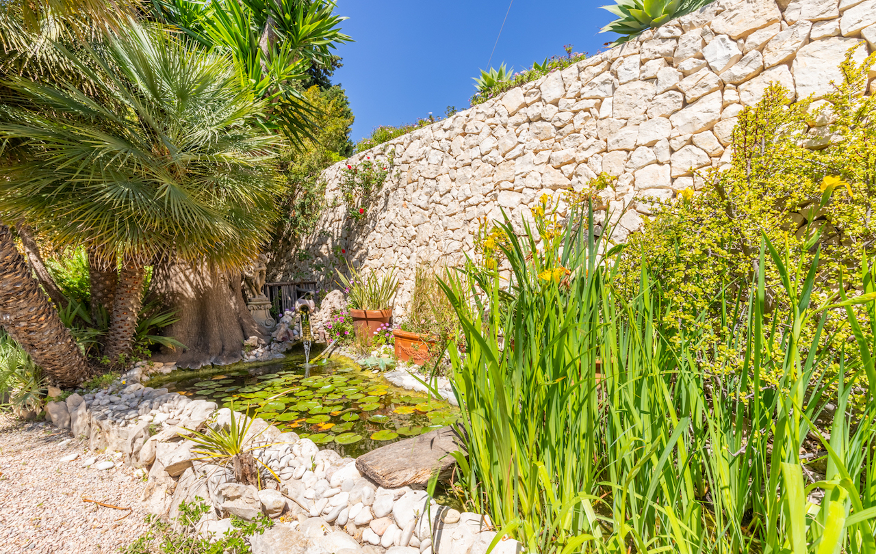 Ornamental pond with water lilies and a small waterfall, bordered by lush Mediterranean plants and a traditional dry stone wall.