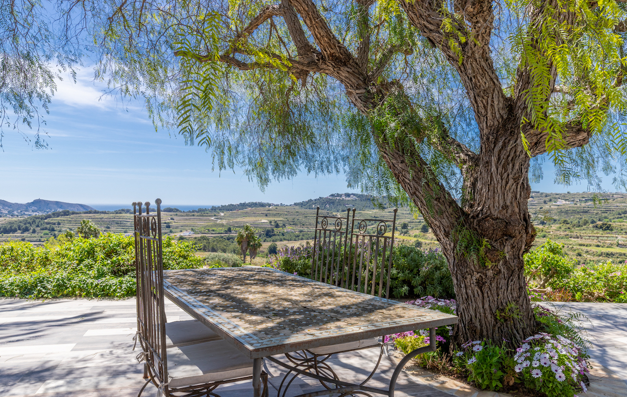Outdoor terrace featuring a mosaic dining table under a mature shade tree, overlooking the rolling vineyards and the Mediterranean Sea toward Moraira.