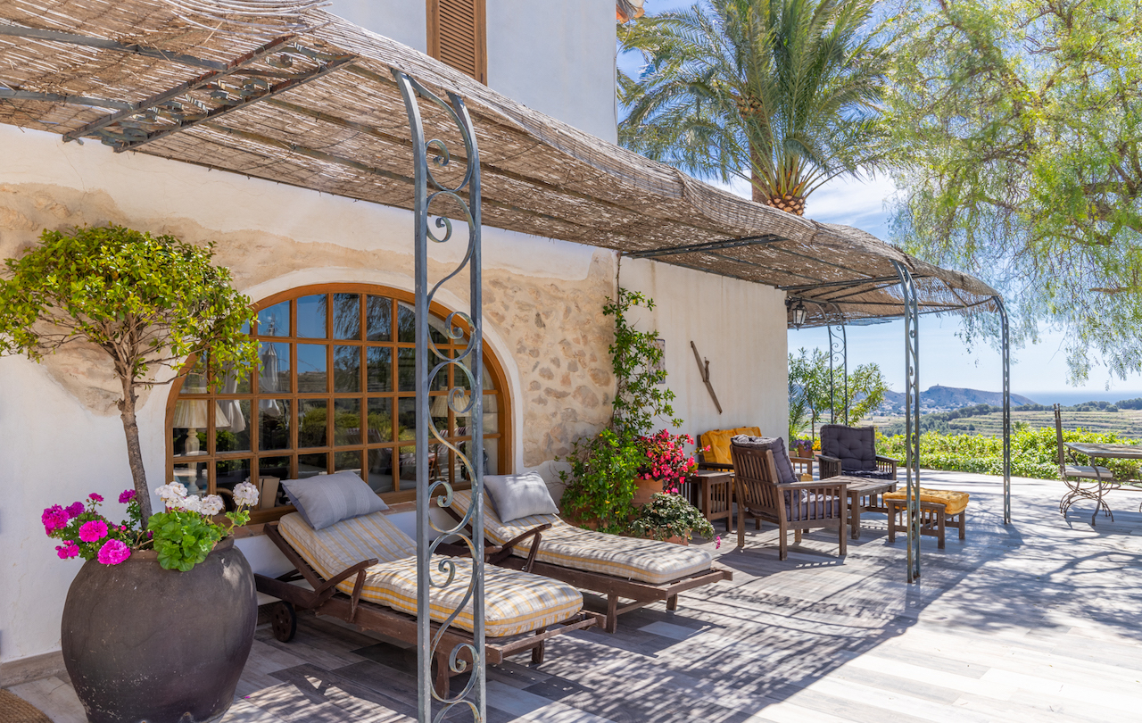 Outdoor terrace featuring a reed-covered wrought iron pergola, sun loungers by a large arched window, and panoramic views of the Mediterranean landscape.