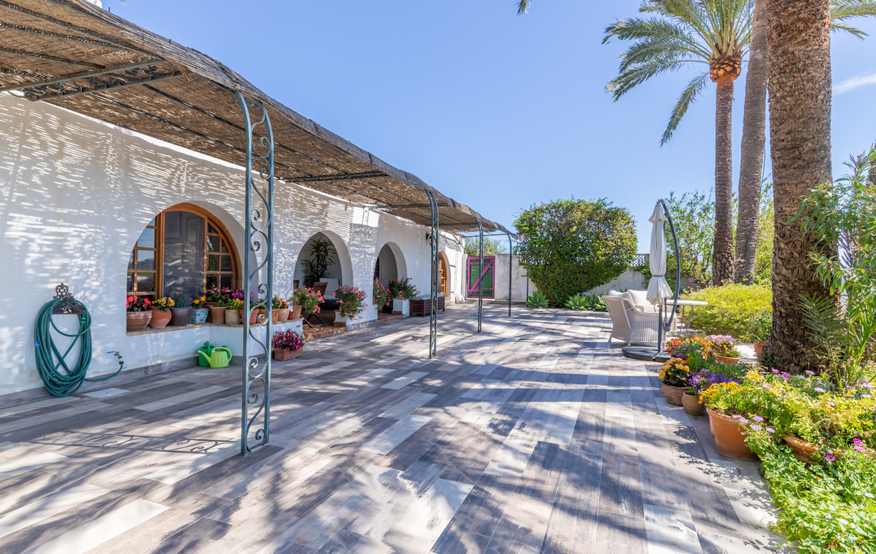 Outdoor terrace featuring wood-effect tiling, a reed pergola supported by wrought iron pillars, and a white facade with traditional arches and potted plants.
