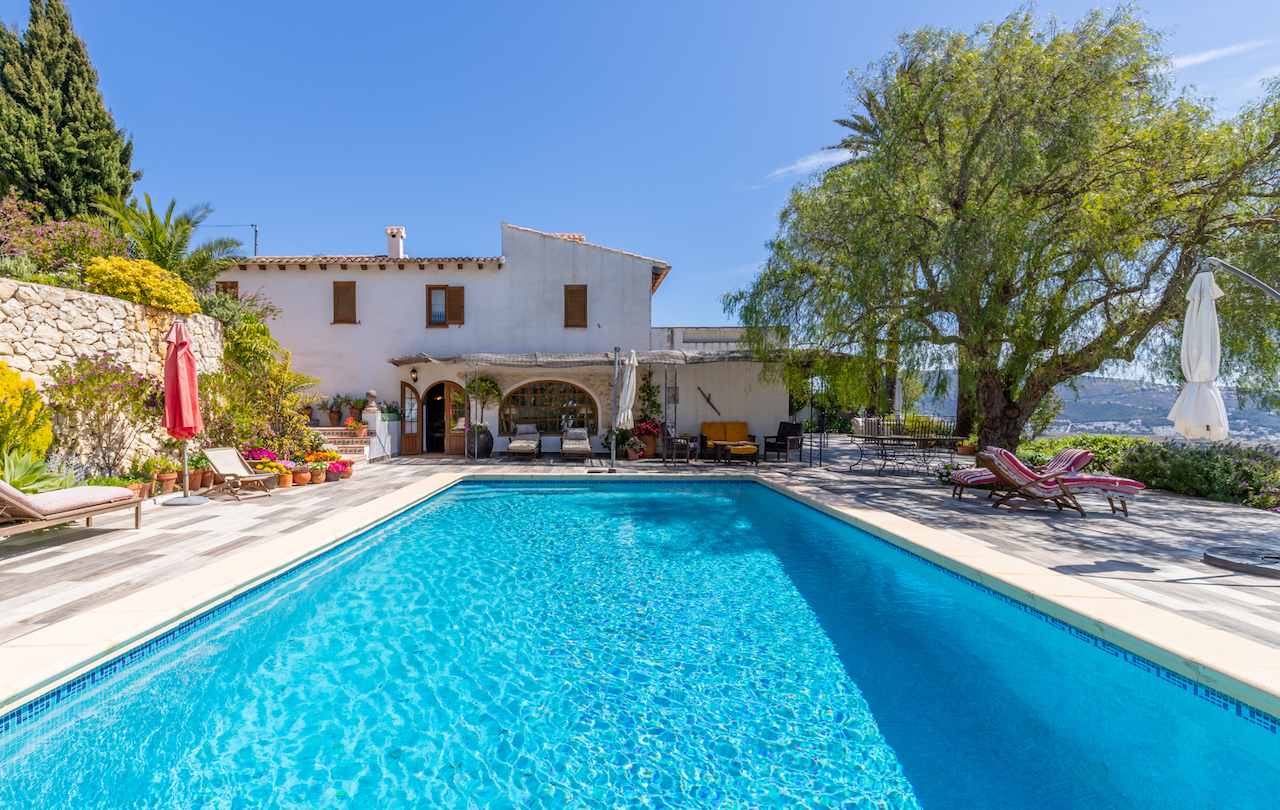 Rectangular swimming pool set within a spacious non-slip tiled terrace, featuring a traditional white facade, stone walls with floral accents, and mature trees.