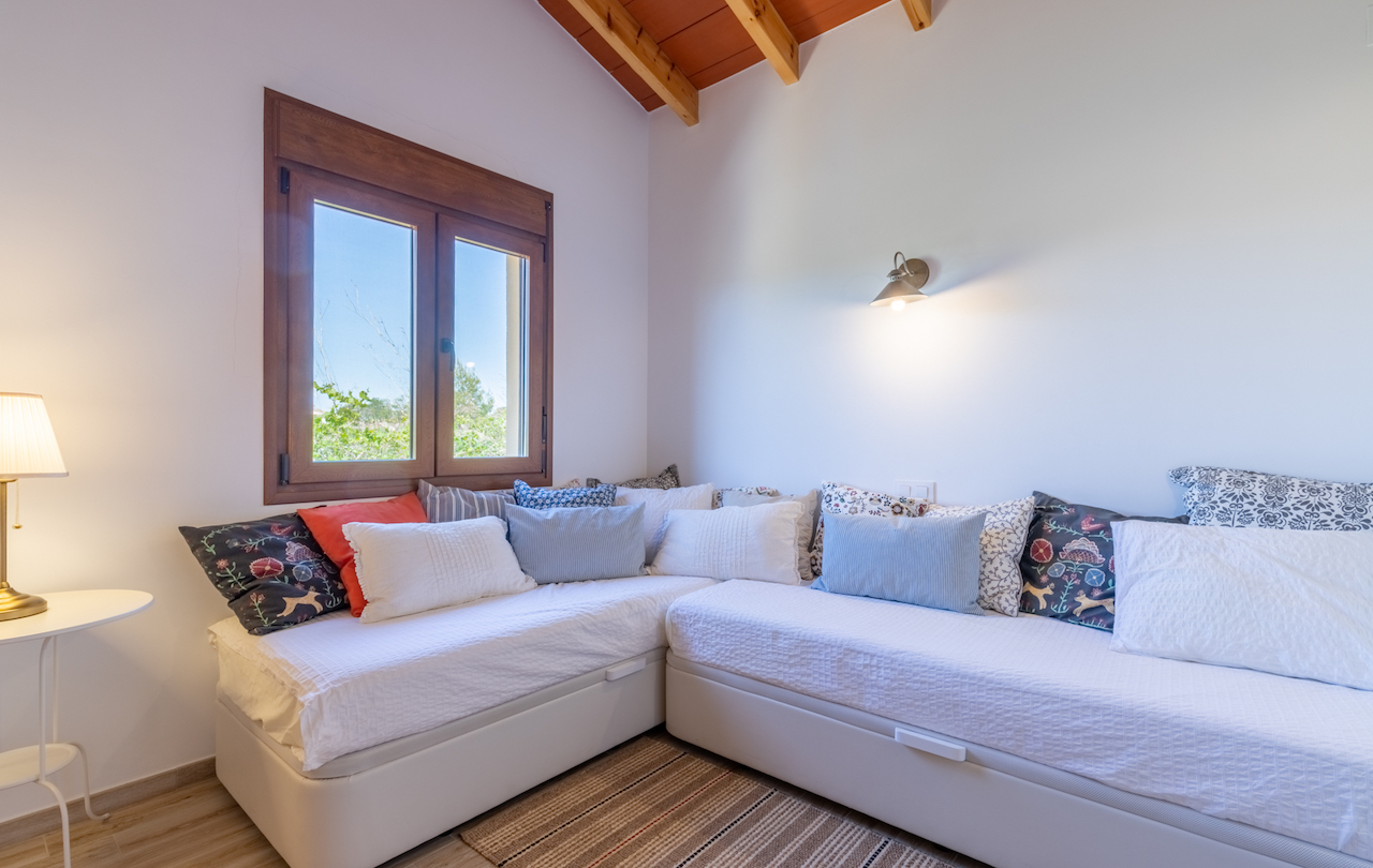 Room featuring an L-shaped daybed with decorative cushions, exposed wooden ceiling beams, and a wooden window framing views of the lush garden landscape.
