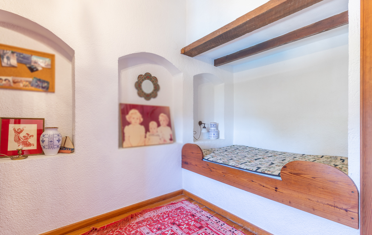 Rustic bedroom featuring a built-in wooden alcove bed, exposed timber beams, and decorative wall niches set against traditional white textured walls.