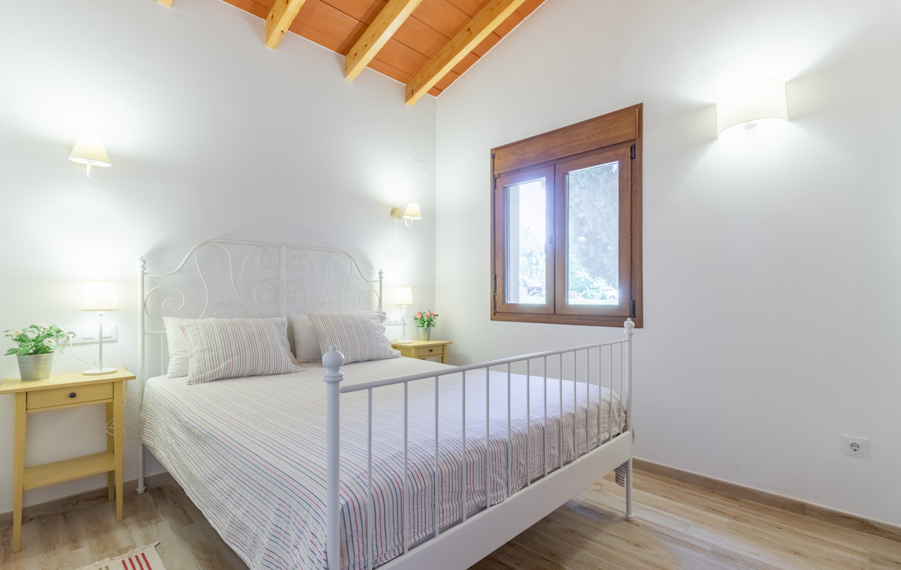 Rustic bedroom featuring a vaulted ceiling with wooden beams, a white wrought iron bed frame, and a wooden-framed window.