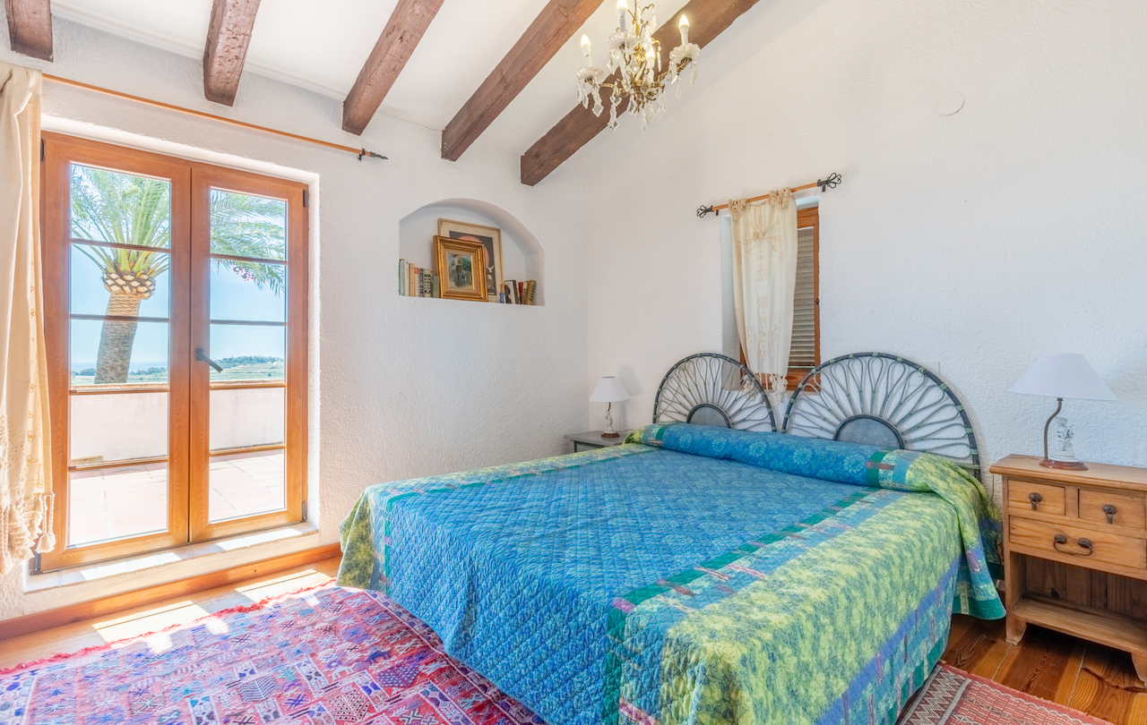 Rustic bedroom featuring exposed wooden beams, a decorative wall niche, and direct terrace access with palm tree views. Includes natural wood flooring and a wrought iron headboard.