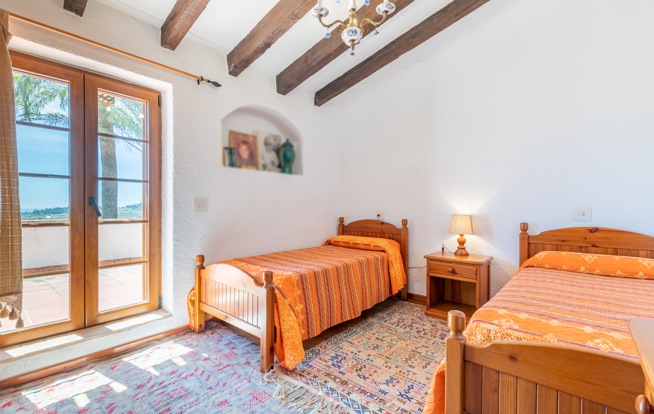 Rustic bedroom featuring exposed wooden beams, twin beds, and a built-in wall niche. Large glass doors open onto a private terrace with views of the Mediterranean landscape.