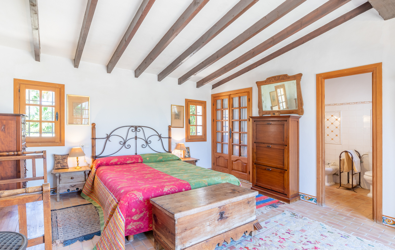 Rustic bedroom featuring exposed wooden ceiling beams, terracotta tile flooring, and traditional furniture. The space includes an integrated en-suite bathroom.