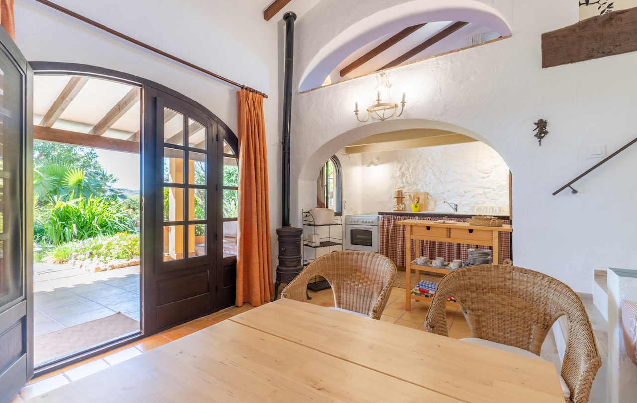 Rustic dining space featuring exposed timber beams, a cast-iron stove, and terracotta tiles. An elegant arch connects the area to a kitchenette with direct access to the garden.