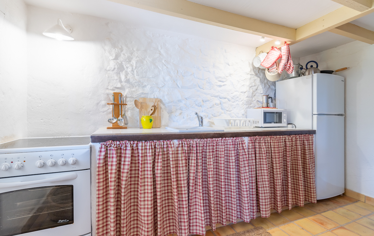 Rustic kitchen featuring white-washed stone walls, exposed wooden beams, and terracotta flooring. Equipped with white appliances and a tiled countertop with classic checkered curtains.